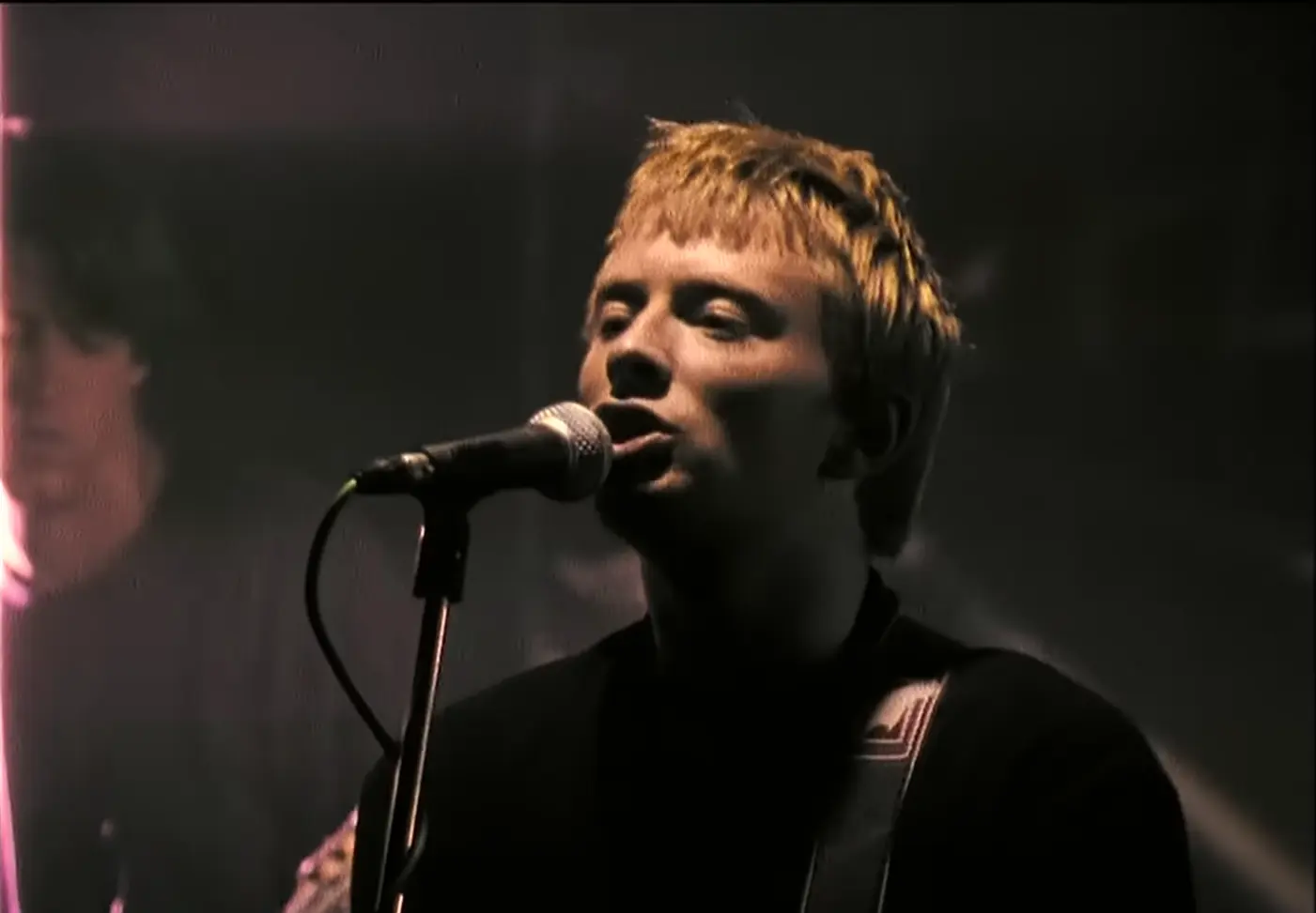 Thom Yorke performing in Radiohead's 'Creep' music video – featuring the band's lead singer with short blonde hair, singing passionately into a microphone under dim, moody stage lighting, holding an electric guitar. Iconic 1990s alternative rock visual style.